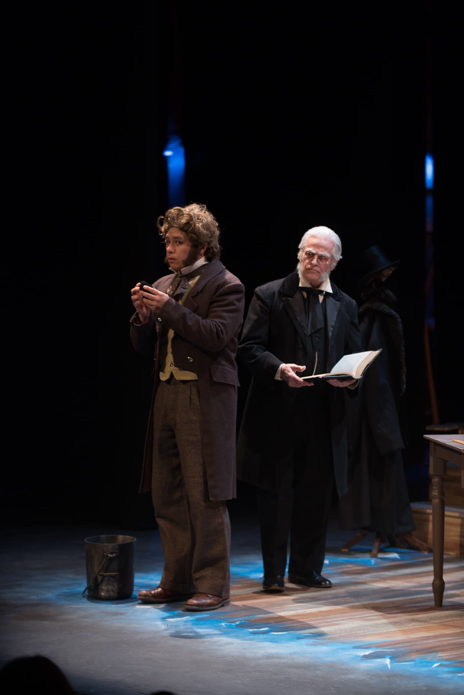 Two men are on stage and dressed in turn of the century clothing. One is wearing a tuxedo while the other is wearing less
expensive clothing.