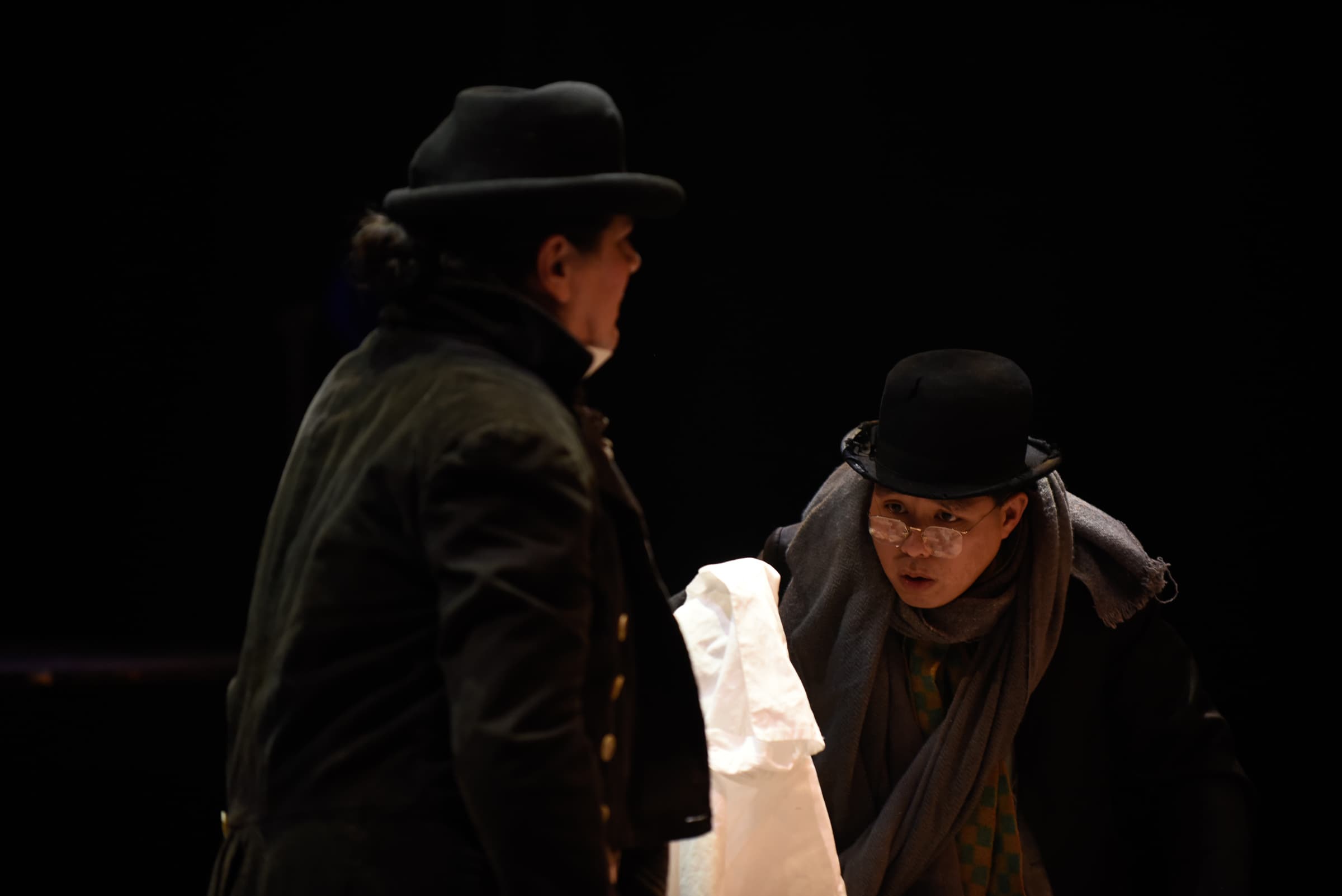 A man is on staged and dressed in turn of the century clothing. He is peering down at something on the lapel of the person
in front of him.