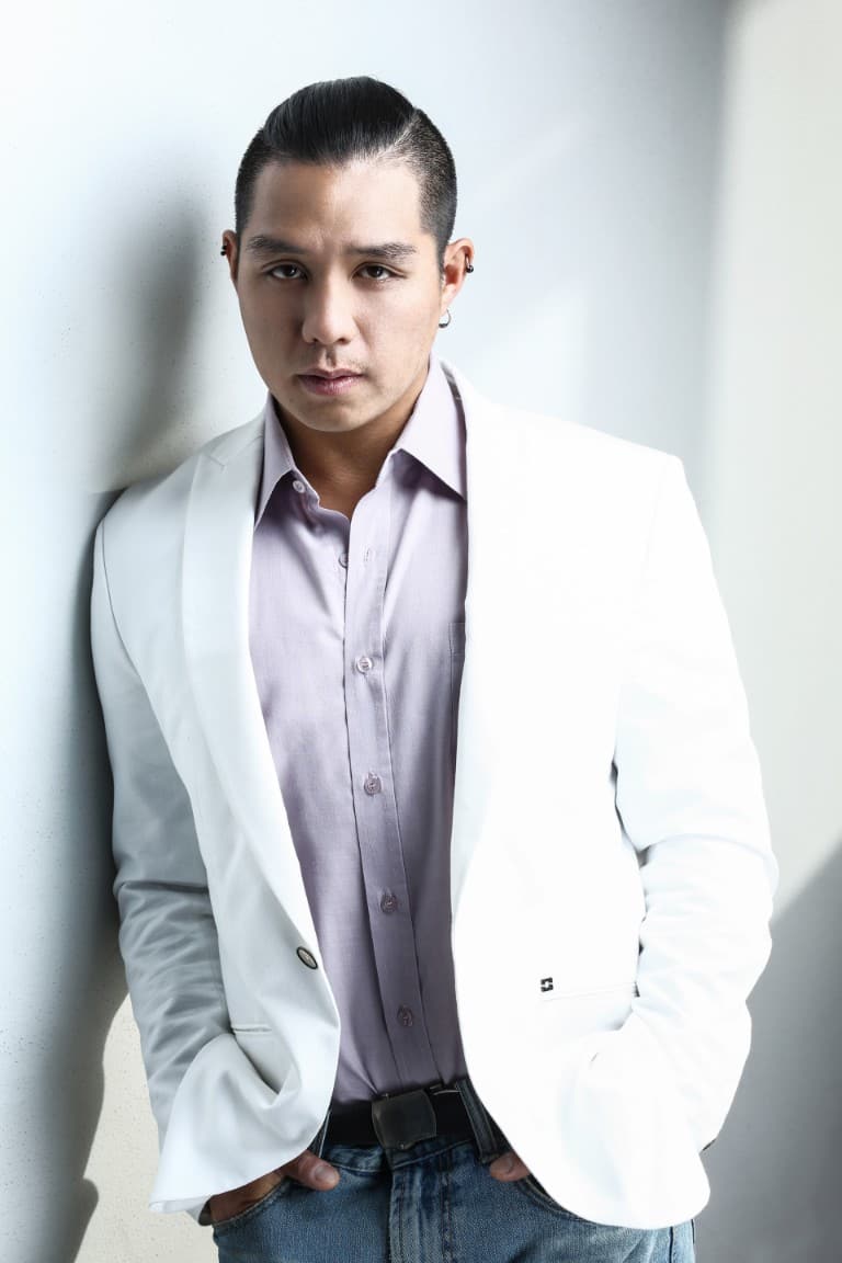 Aeryhn wearing a white blazer over a lilac shirt, leaning on a wall and looking at the camera.