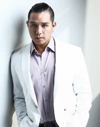 Aeryhn wearing a white blazer over a lilac shirt, leaning on a wall and looking at the camera.