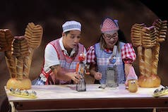 A man and a woman are standing behind a desk and using kitchen utensils as puppets. A hand mixer, a cheese grater, and a potato have clay heads on them and they're being moved around by the man the woman.