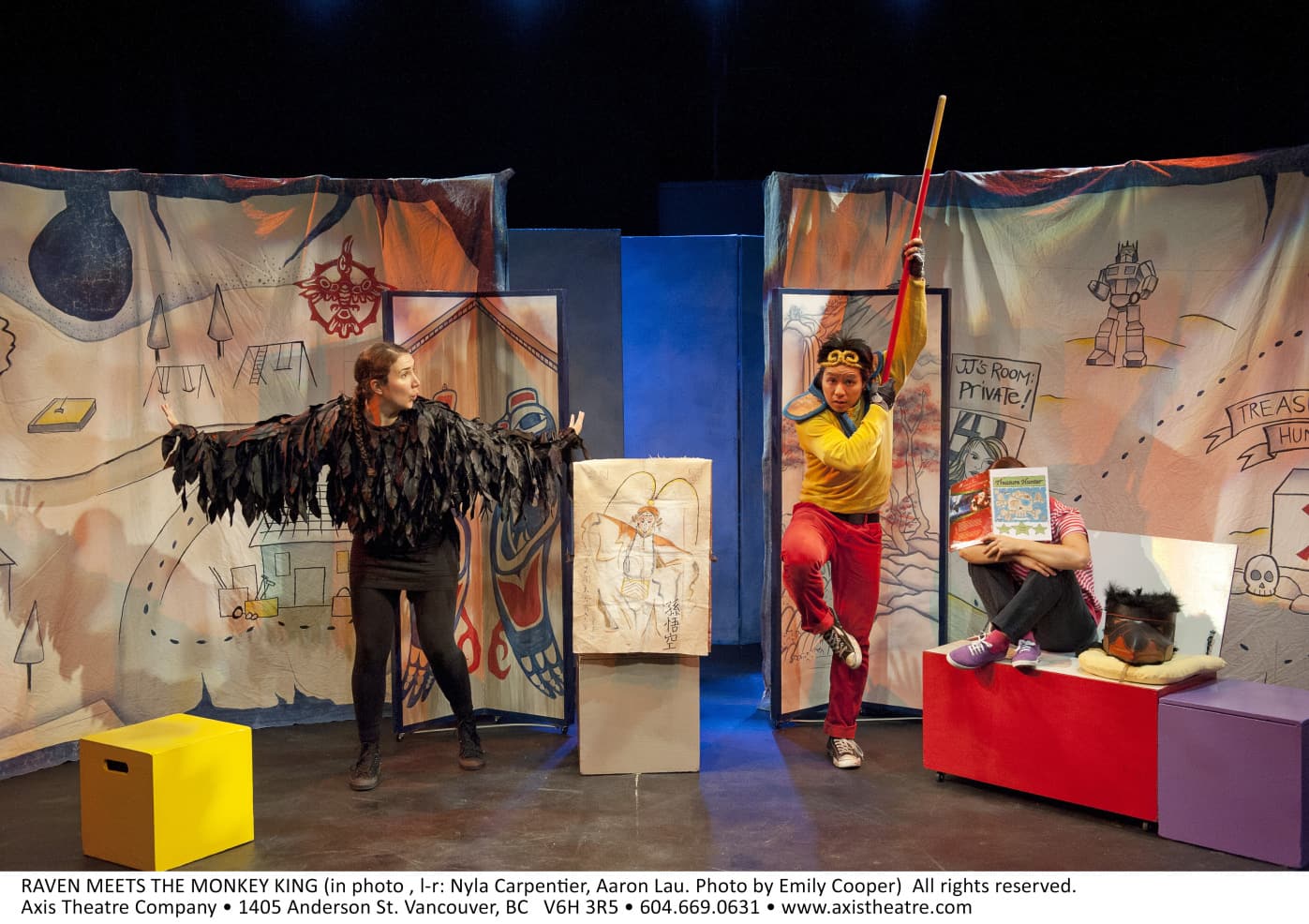 Three people are on stage, one dressed as a raven, one as a monkey, and another is covered their face with a book.