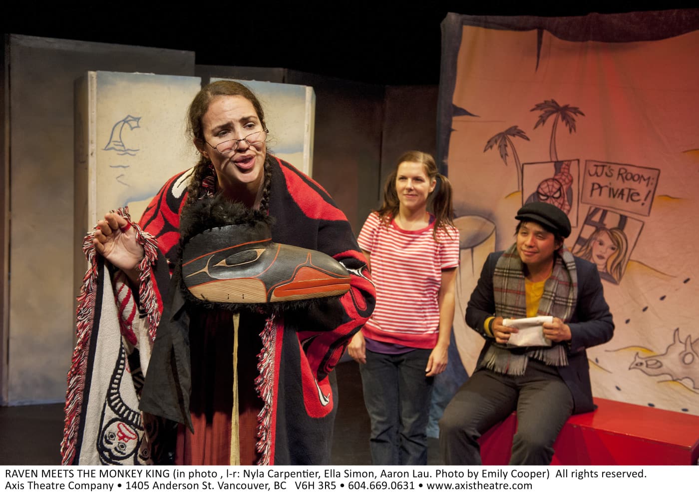 A woman wears traditional First Nations patterns from Western Canada while holding a wooden mask carved in the shape of a
raven. Two people watch her from the back.
